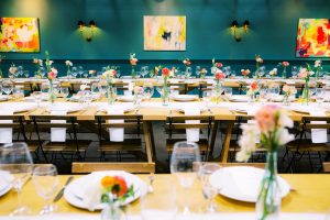 Mid-century modern event venue interior with emerald green furniture, stained glass windows, and an open view to a courtyard decorated with string lights at sunset.